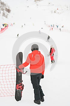Man in black helmet wearing winter sportswear holding snowboard looking at snow mountain with skiers and snowboarder young girl in