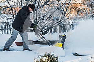 A man in a black jacket and a gray pants is brushing white snow with the yellow electric snow thrower in winter