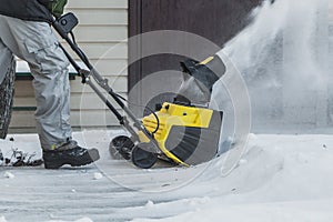 A man in a black jacket and a gray pants is brushing white snow with the yellow electric snow thrower in winter