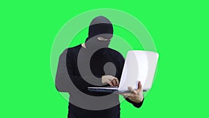 A man in a black mask enters data into a laptop, on a green screen background