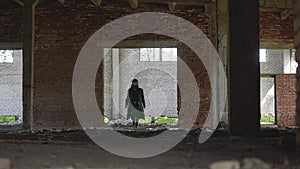 A man in a black raincoat, hat and black glasses, with a beard and dreadlocks is a large room abandoned factory.