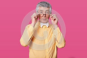 Man with black rounded glasses on tip of nose. Suprised old male holds spectacles with hands. Eldery senior with wide opened eyes