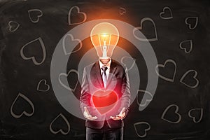 Man in black suit with light bulb instead head, levitating red glaring heart with his palms, standing against black