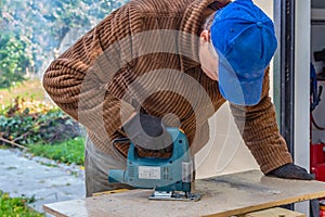 A man in the black working gloves and a brown jacket and a blue hat cuts a board using a Jigsaw power tool on a work