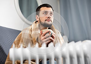 Man with a blanket on his shoulders with cup near electric heater