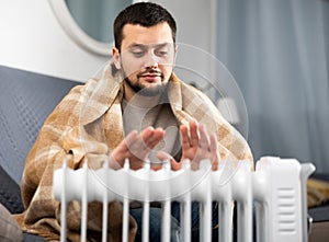 Man with a blanket on his shoulders with cup near electric heater