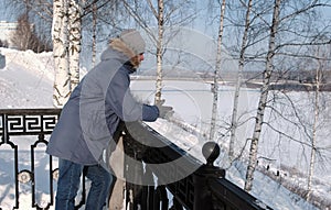 Man in blue down jacket with fur hood stands at the fence And look around in the winter Park.