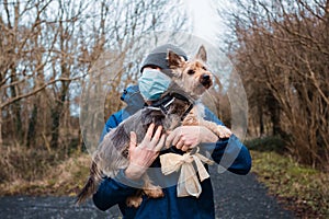 Man in blue jacket and grey hat and medical mask hugging yorkshire terrier in a park. Expression of care and sentiment to your