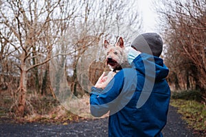 Man in blue jacket and grey hat and medical mask hugging yorkshire terrier in a park. Expression of care and sentiment to your