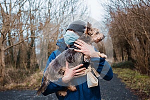 Man in blue jacket and grey hat and medical mask hugging yorkshire terrier in a park. Expression of care and sentiment to your