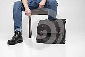 Man in a blue jeans and black boots sits on a chair with a brown men& x27;s shoulder leather bag for a documents and laptop