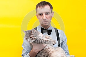 Man in blue shirt, black suspender and bow tie holding grey striped tabby cat with outstretched paws and looking at camera