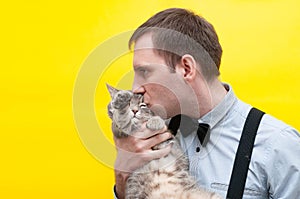 Man in blue shirt, black suspender and bow tie kissing grey striped tabby cat with outstretched paws