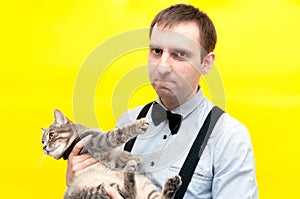 Man in blue shirt with long sleeves, bow tie and suspender holding cute grey tabby cat  and looking at camera