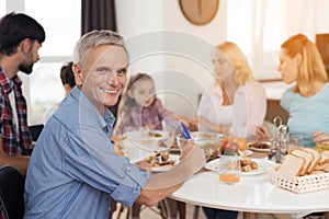 A man in a blue shirt is sitting at a table and posing against the background of his family, who is sitting and eating