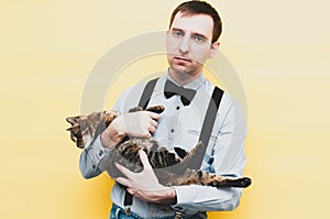 Man in blue shirt, suspender and bow tie holding cute brown tabby cat on back and looking at camera