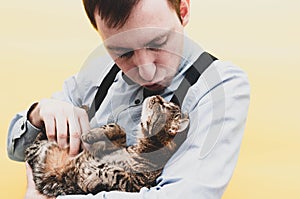 Man in blue shirt and suspender holding and petting cute brown tabby cat on back and showing air kiss