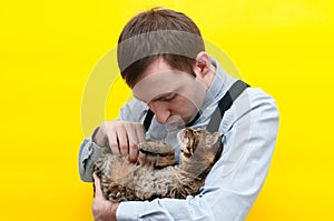 Man in blue shirt and suspender holding and petting cute brown tabby cat on back and showing air kiss