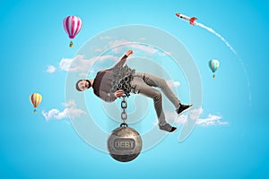 Man in blue sky bound with chains to big metal ball that reads `DEBT` and is pulling him down.