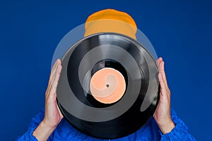 man in a blue sport 90s style jacket and yellow hat holding vinyl record, man hides his face under vinyl disc