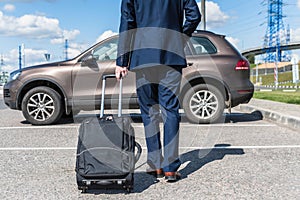 Man in a blue suit with a suitcase at the airport parking on the backdrop of his car. Business trip concept.