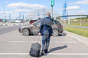 Man in a blue suit with a suitcase walking to his car and calling by phone at the airport parking. Business trip concept.