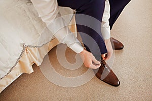 A man in a blue suit ties up shoelaces on brown leather shoes brogues on a wooden parquet background