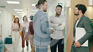 A man in a blue suit walks and joins a pair of workers as they are having a conversation in the hall, two working women