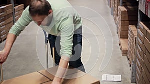 A man in a blue sweater is taking boxes from the shelf, putting them on the trolley in a storage warehouse