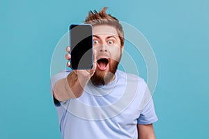 Man in blue T-shirt holding out cellphone and looking at camera with big eyes and open mouth.