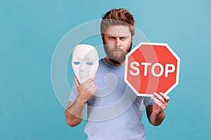 Man in blue T-shirt holding red stop sign and white mask, looking at camera with strict expression.