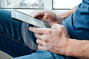 A man in a blue t-shirt and jeans holds an electronic book in his hands. Reading books at home by the window