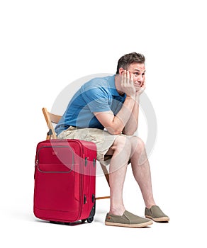 Man in a blue T-shirt and white shorts sits on a chair near a red suitcase, waiting. Isolated on white background
