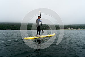 Man in blue wetsuit is paddling on a yellow SUP board on a sea waves at cloudy day.