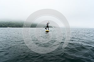 Man in blue wetsuit is paddling on a yellow SUP board on a sea waves at cloudy day.