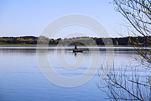 A man on a boat fishing in vast blue lake water with lush green trees and plants on the banks of the lake with blue sky