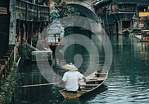 Man in a boat with a traditional triangular chinese hat standing in waters of Tuo river flowing through the centre of ancient city