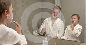 Man and boy in bathroom putting shaving cream on face, child son with dad in bathrobes