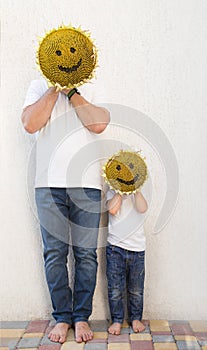 Man and boy holding yellow sunflower head in front of faces, on which muzzle with smile is drawn