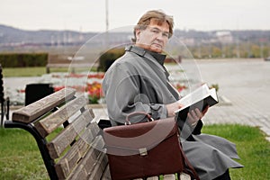 A man with a briefcase, looking like a writer or a preacher, is reading a book sitting on a bench on a city street