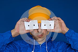 man in a bright blue jacket and yellow hat holding vintage audio cassettes on a bright blue background