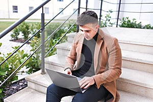 Man in a brown coat with a laptop in his hands sits on the stairs. Student works at the computer
