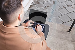 Man in a brown coat with a laptop in his hands sits on the stairs. Student works at the computer