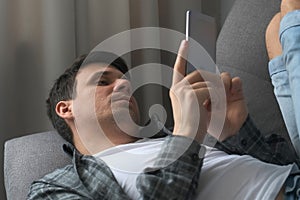 Man is browsing smartphone finding a woman on dating site online lying on sofa.