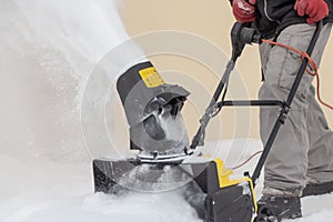 Man is brushing white snow with the yellow electric snow thrower in a winter garden