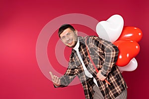 A man with a bunch of red and white balloons smiling, extending hand to the camera ove red background with blank space.