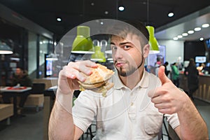 A man with a burger in his hands sits in a fast-food restaurant and shows a thumbs up. A man likes a burger
