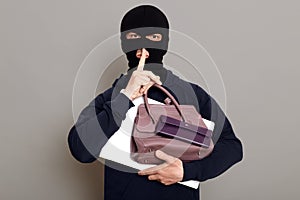 Man burglar holds laptop, wallet and woman`s purse in his hands, holds finger to his mouth, shows gesture quietly, the robber