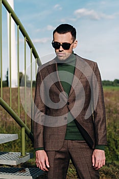 A man in a business suit with a green Golf shirt stands next to a windmill against the background of the field and the blue sky.