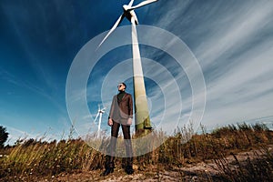 A man in a business suit with a green Golf shirt stands next to a windmill against the background of the field and the blue sky.